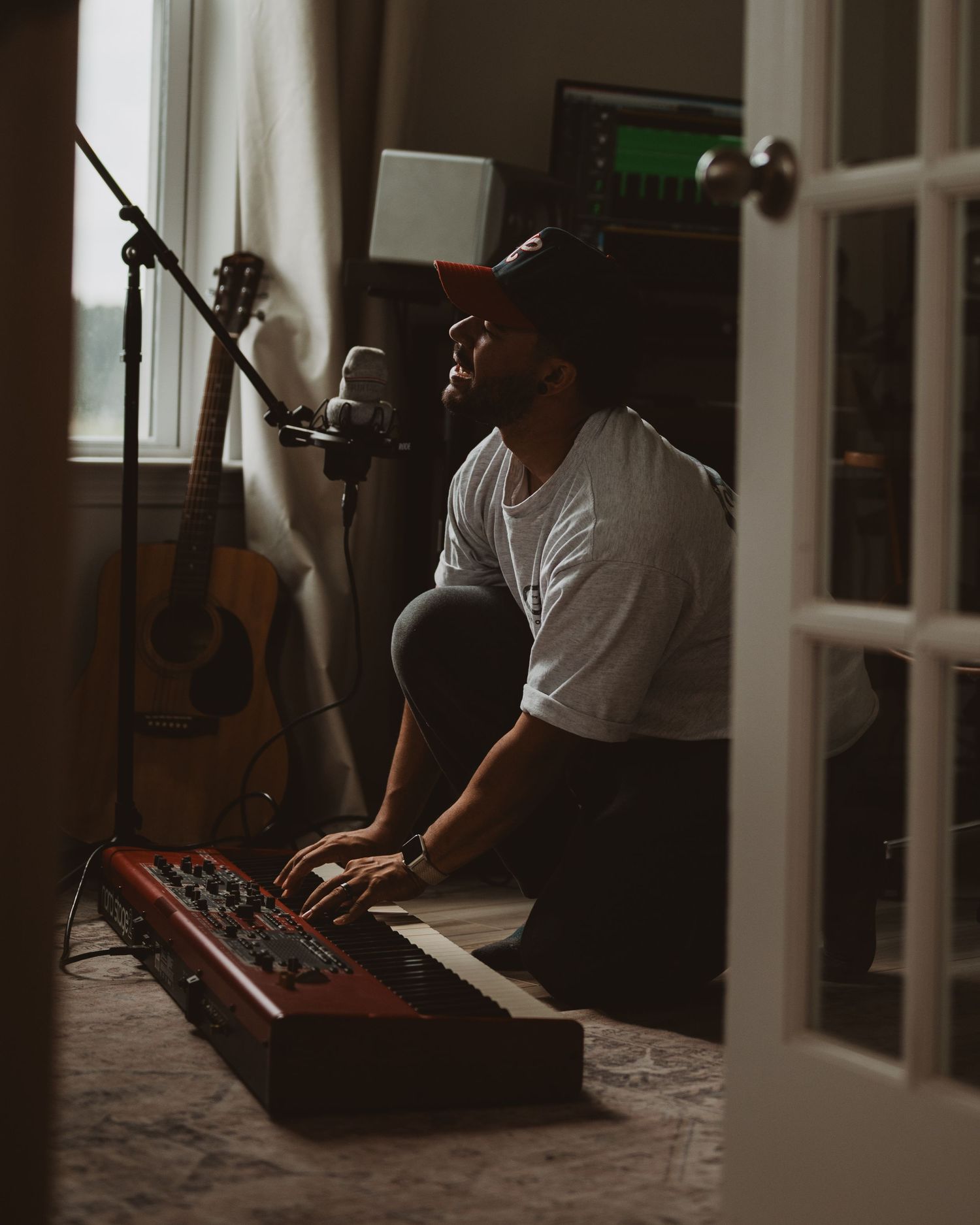 A musician with wireless headphones performs at a home studio setup with microphone and keyboard in moody lighting.