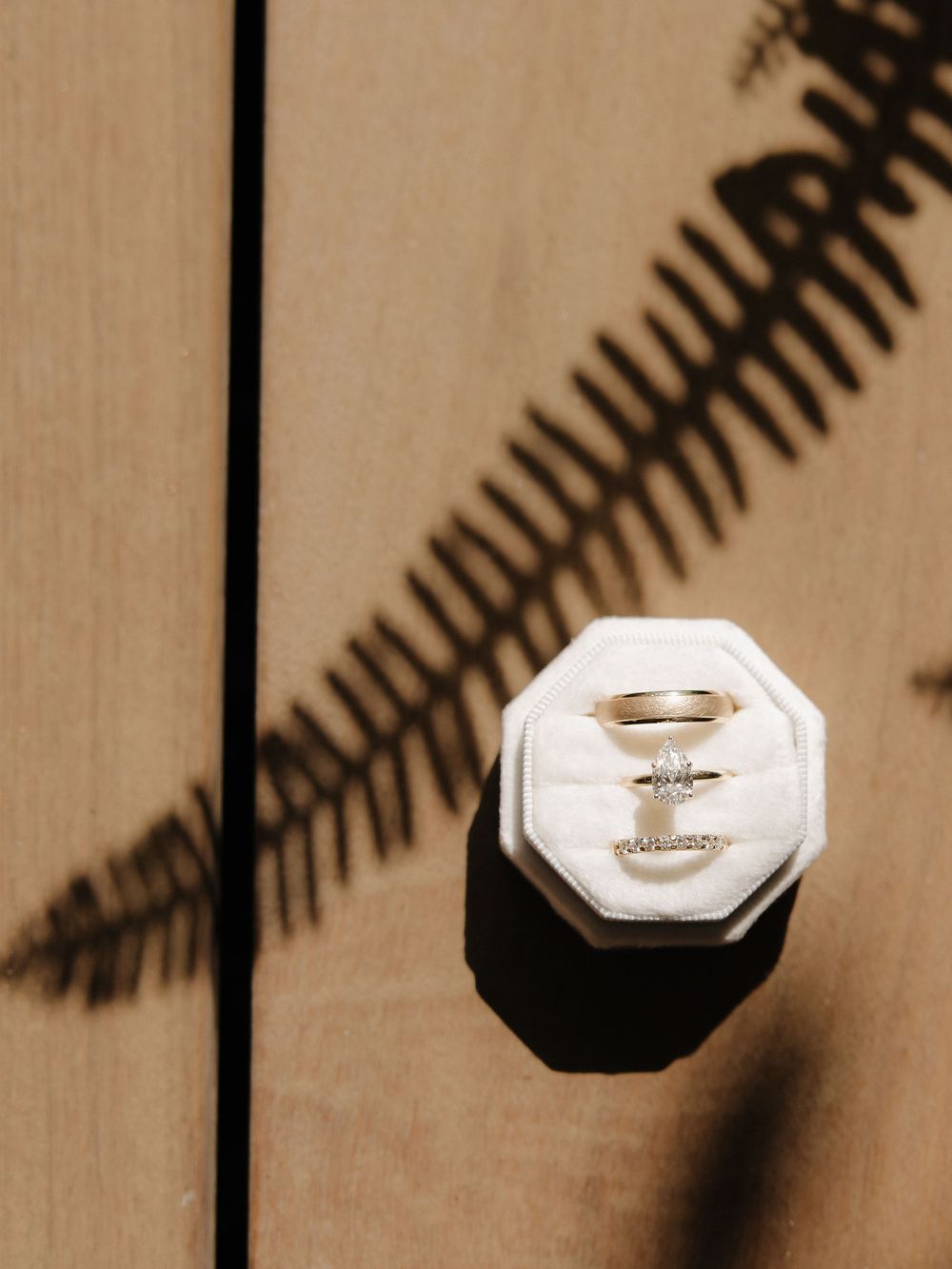 Wedding ring in a white octagonal box casting a palm frond shadow on a wooden surface.
