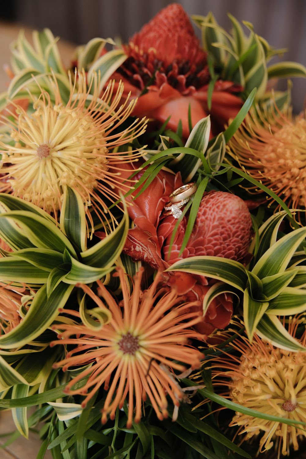 Exotic tropical floral arrangement featuring orange pin cushion proteas and red flowers with green foliage.