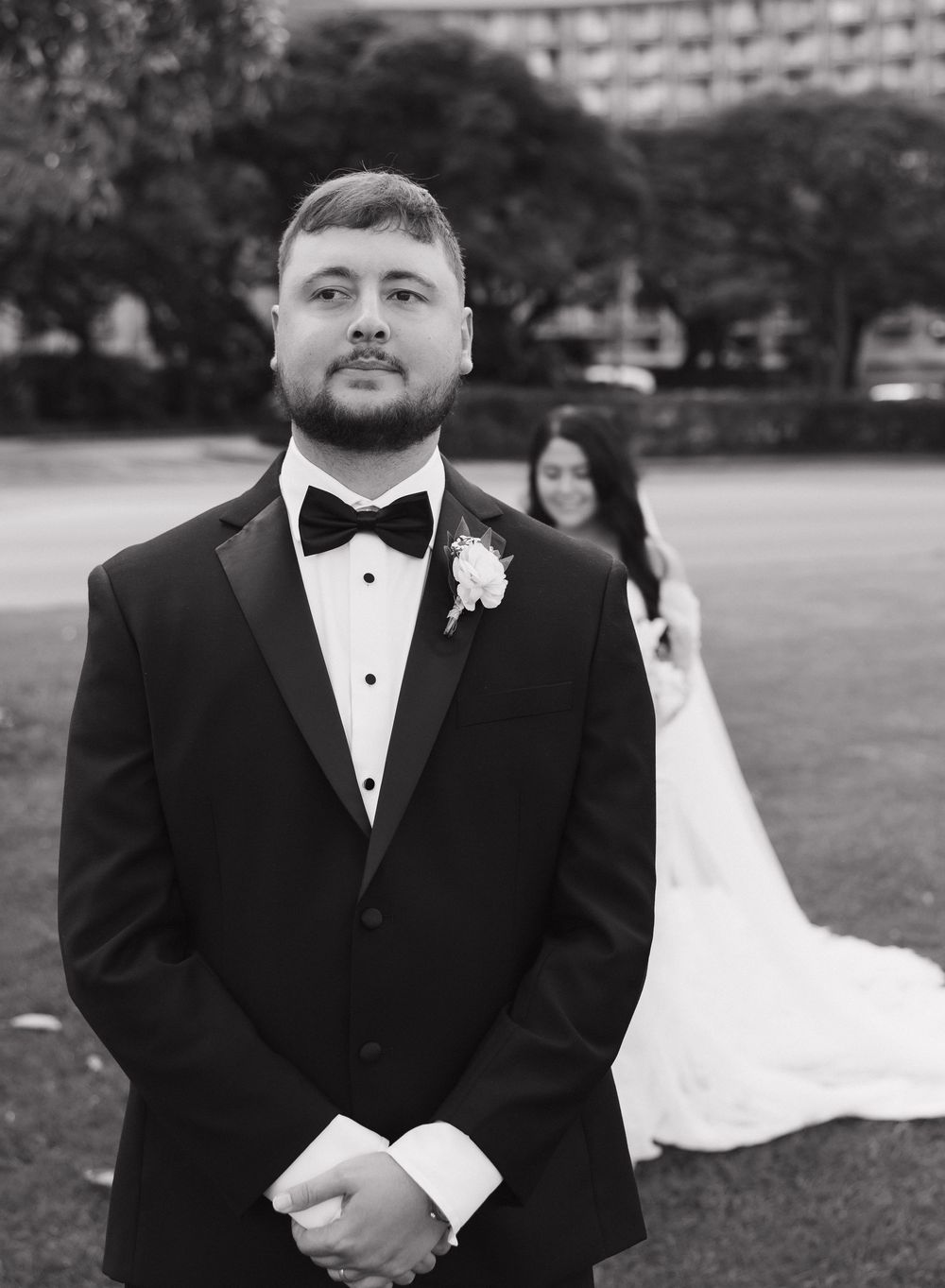 A groom in classic tuxedo poses in a garden while his bride stands in the background.