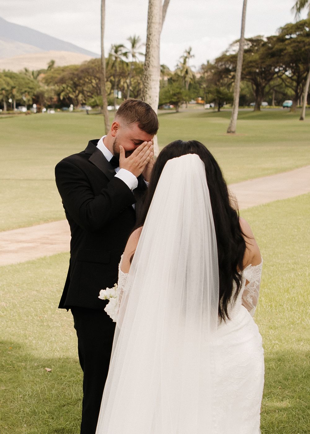 An emotional wedding moment captured on a lush green lawn with palm trees and mountains in the background.