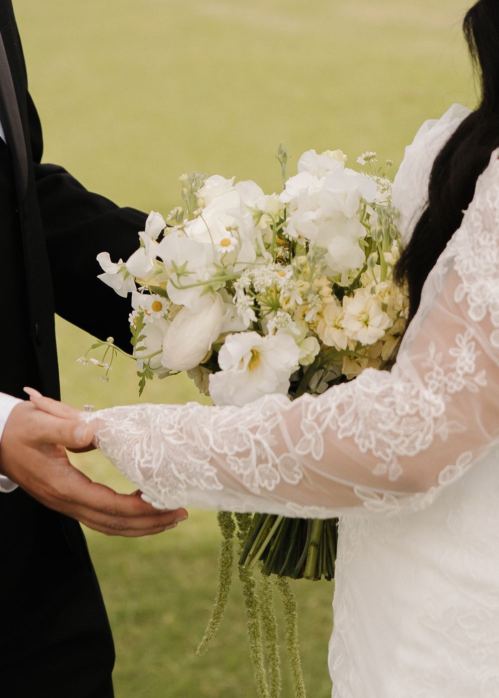 Close up of an elegant white lace wedding bouquet with white roses being held by a bride and groom at an outdoor ceremony.