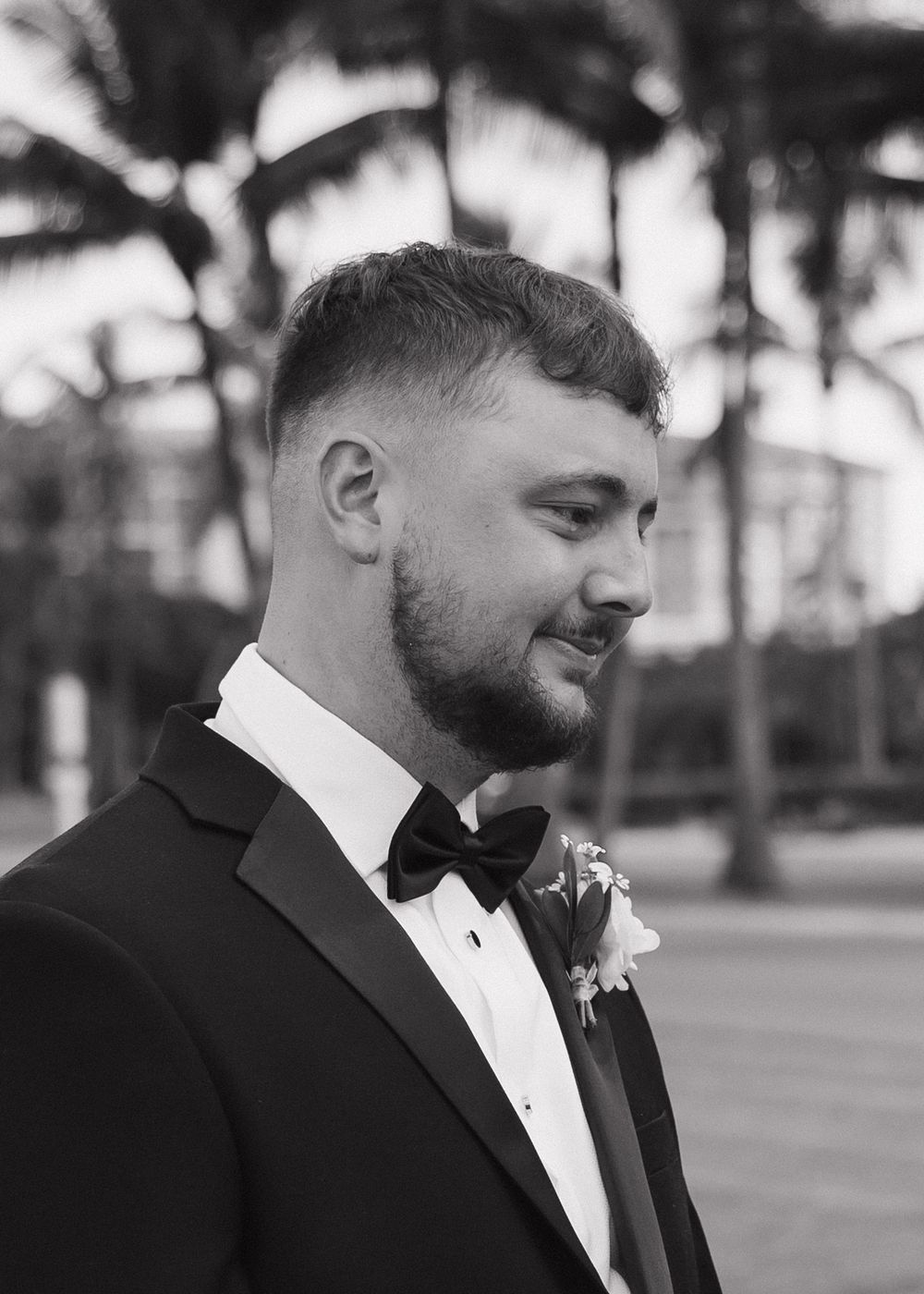 A black and white portrait of a groom in a classic tuxedo with a boutonniere against palm trees.