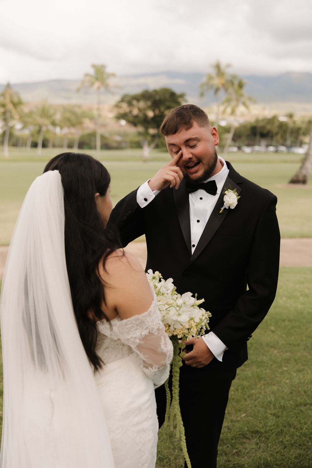 A couple sharing an emotional moment during their wedding ceremony on a palm tree-lined lawn in Hawaii.