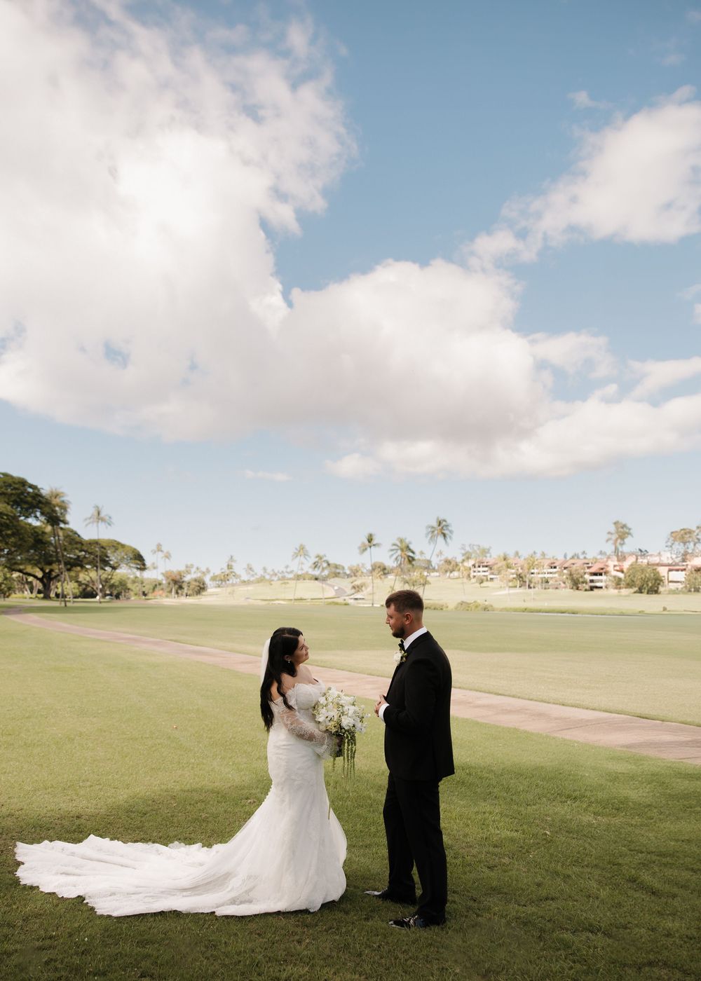 A wedding couple stands together on a lush green field under a bright blue sky.