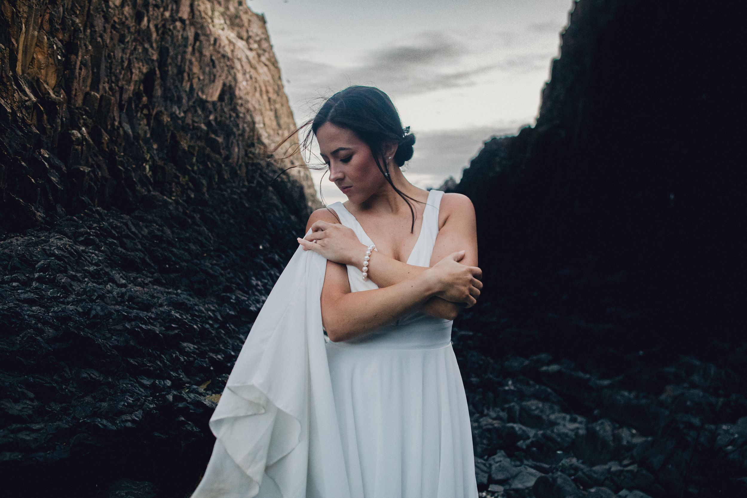 A person in a flowing white dress stands between rocky cliffs at dusk while holding fabric against their body.
