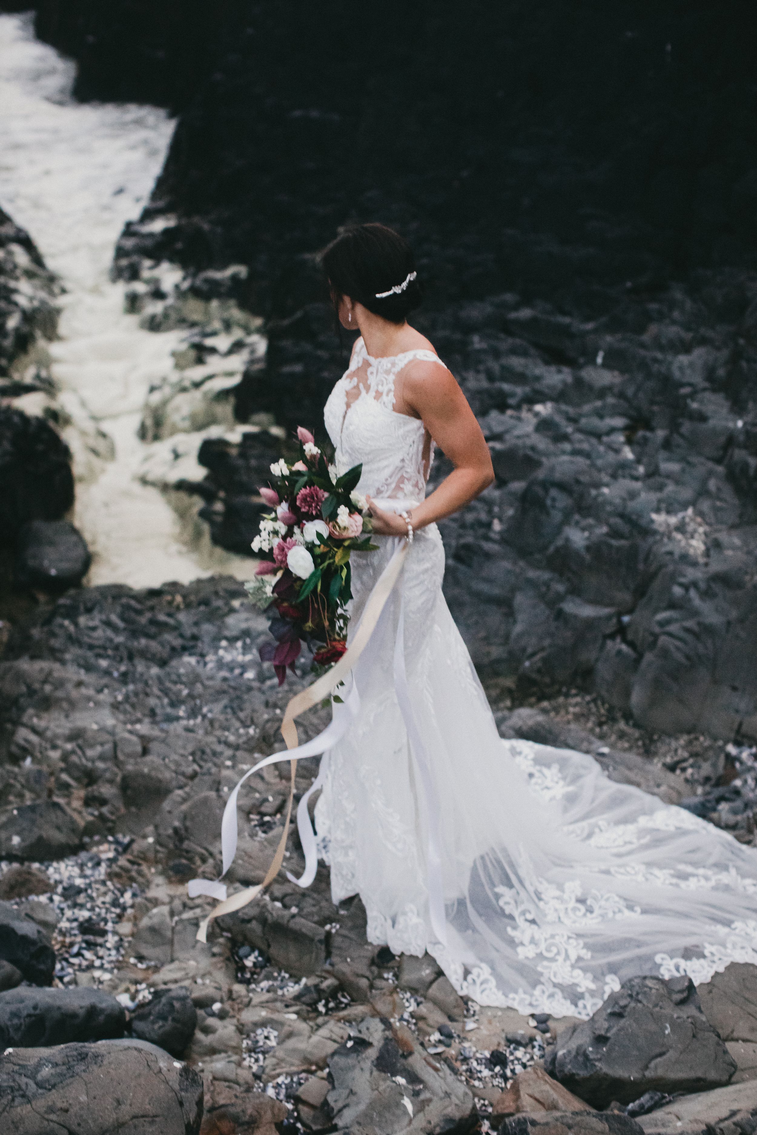 An elegant white lace wedding dress with flowing train photographed against dramatic rocky waterfall backdrop.