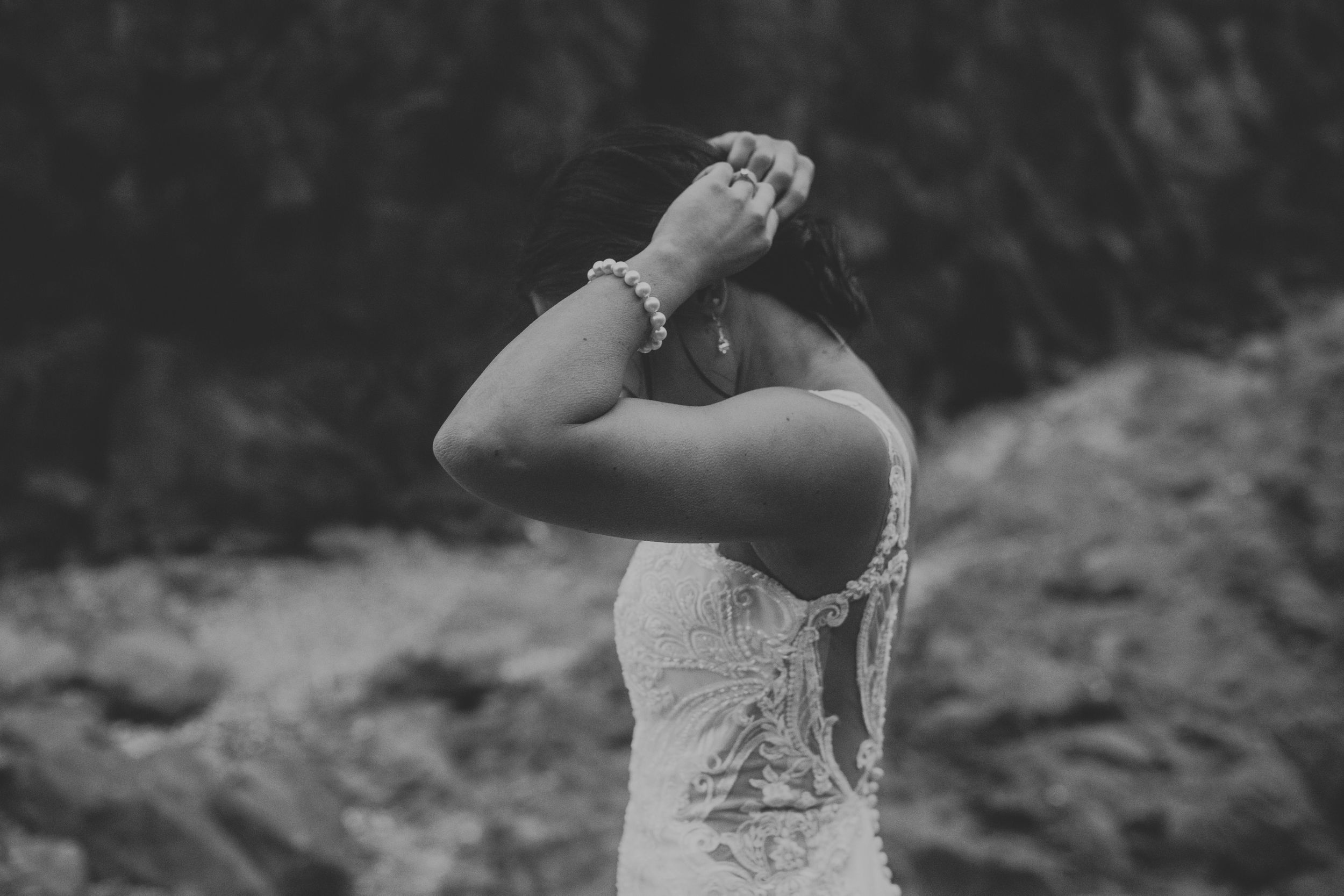 Black and white photo of bride in lace wedding dress holding head while posing outdoors in nature.