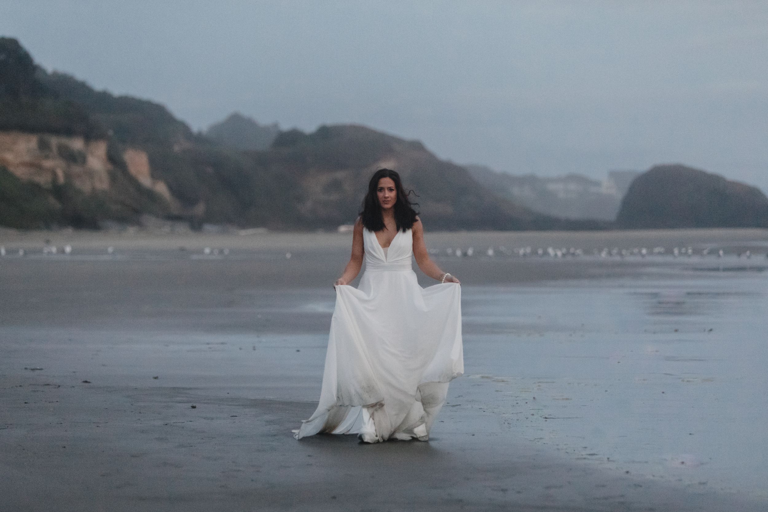 A person in a flowing white wedding dress stands on a misty beach with cliffs in the background at dusk.
