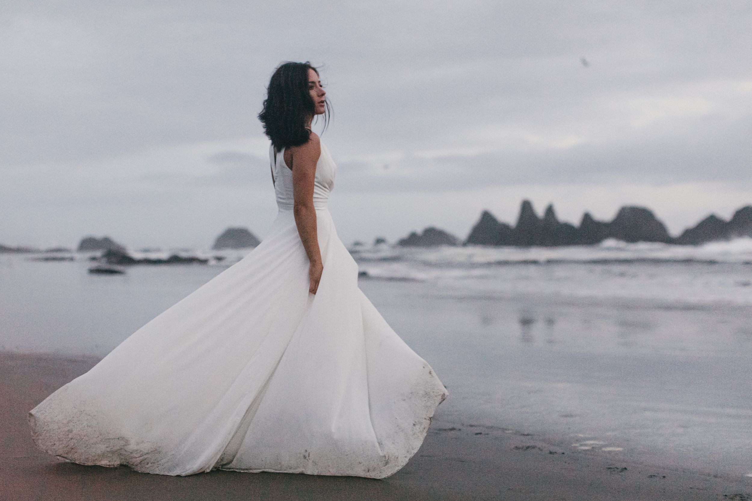 A wedding dress twirls on a moody Pacific Northwest beach with dramatic rock formations in the background.