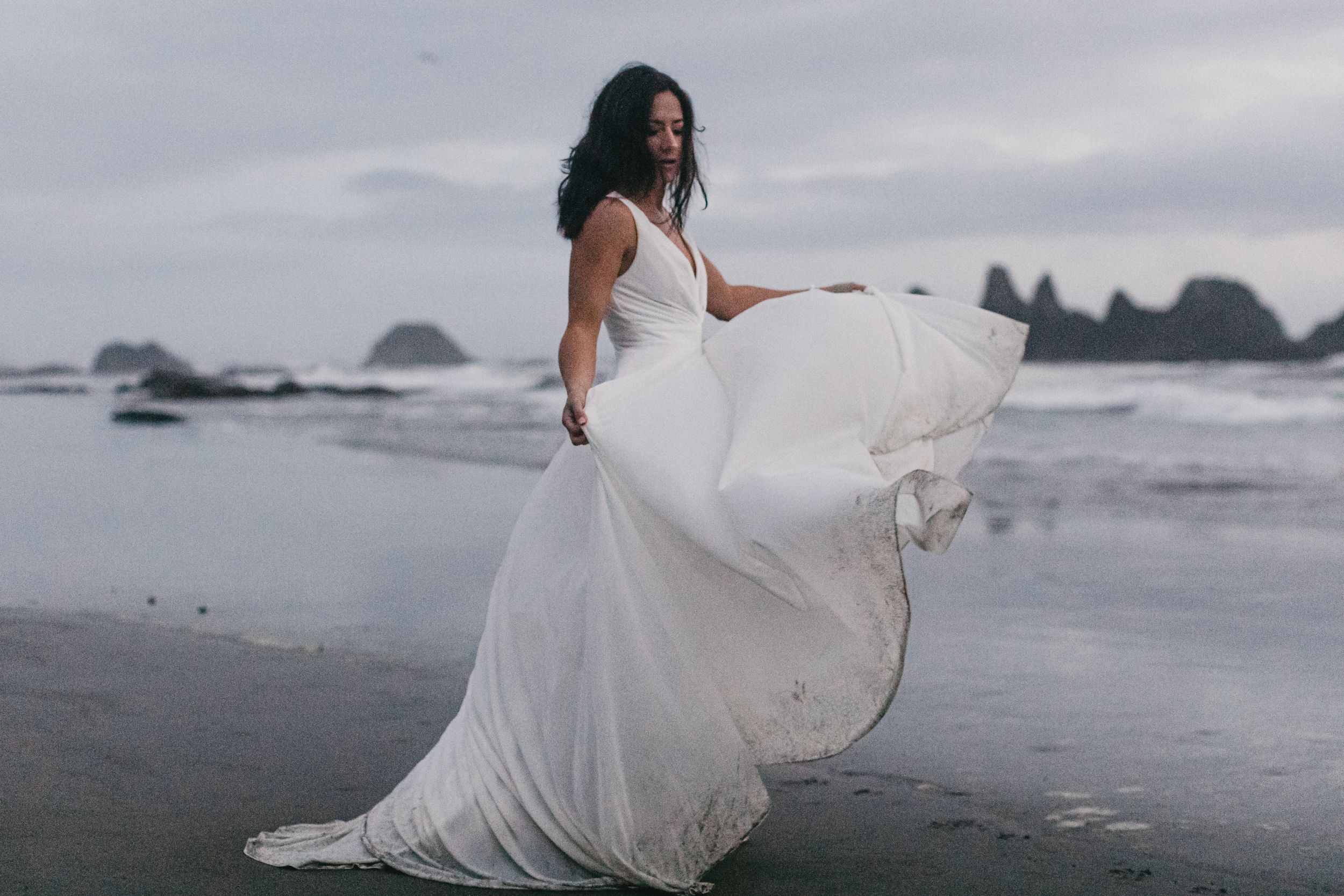 A person in a flowing white wedding dress walks barefoot along a misty rocky beach at sunset.