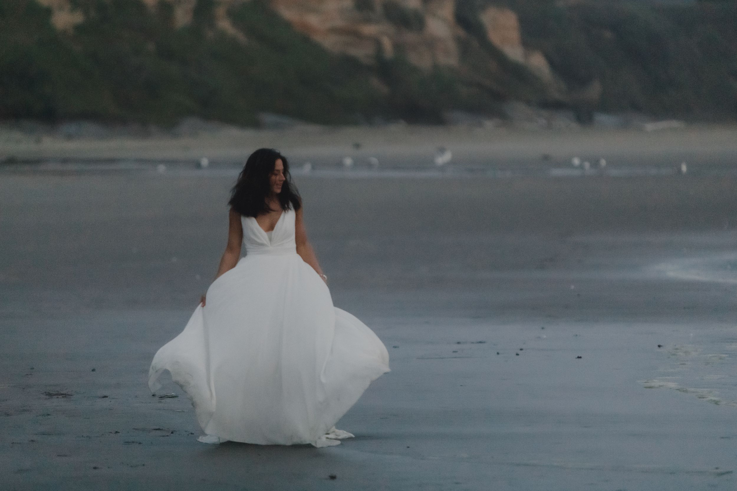 Bride in flowing white dress walks barefoot along misty beach shoreline at sunset.