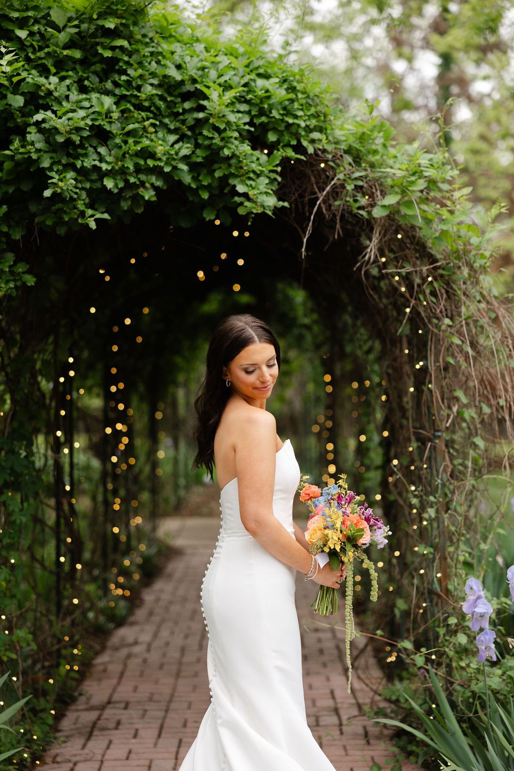 A bride in a white fitted dress holds a colorful bouquet under a garden archway with twinkling lights and greenery. Top 10 Nashville Wedding