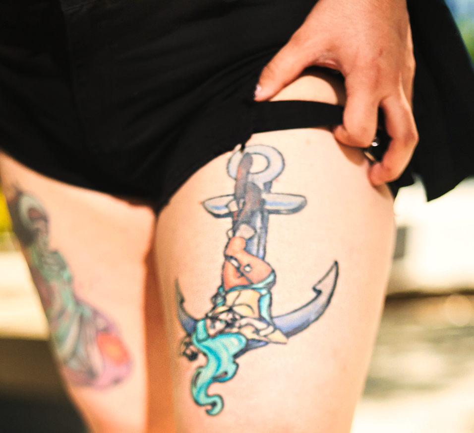 A mermaid sitting on an anchor tattoo design in blue and gray tones on someone's thigh.
