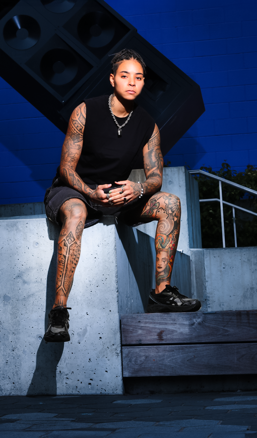 Person in black tank top with tattoos sitting on concrete steps against a blue background.