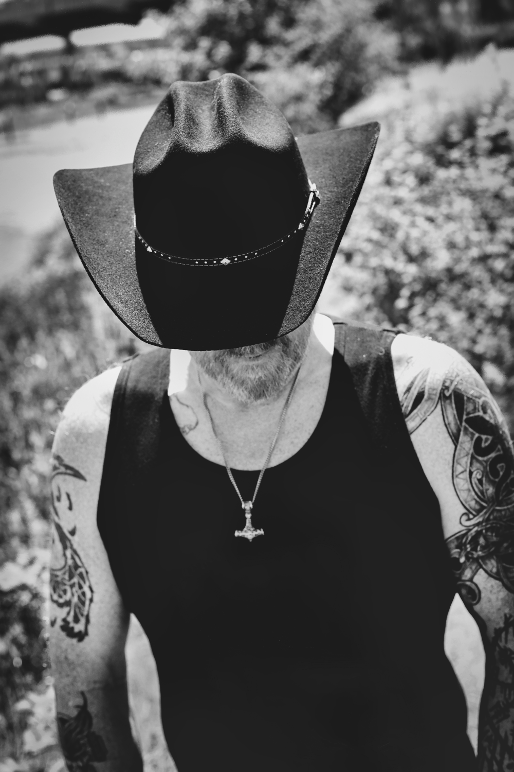 Black and white portrait of person in cowboy hat with tattoos wearing tank top and cross necklace.