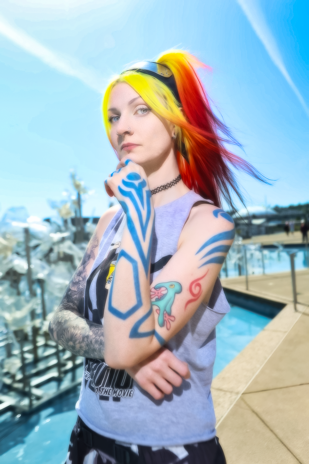 Cosplay photograph showing a person with vibrant yellow and red hair wearing a gray tank top with visible tattoos by a marina.