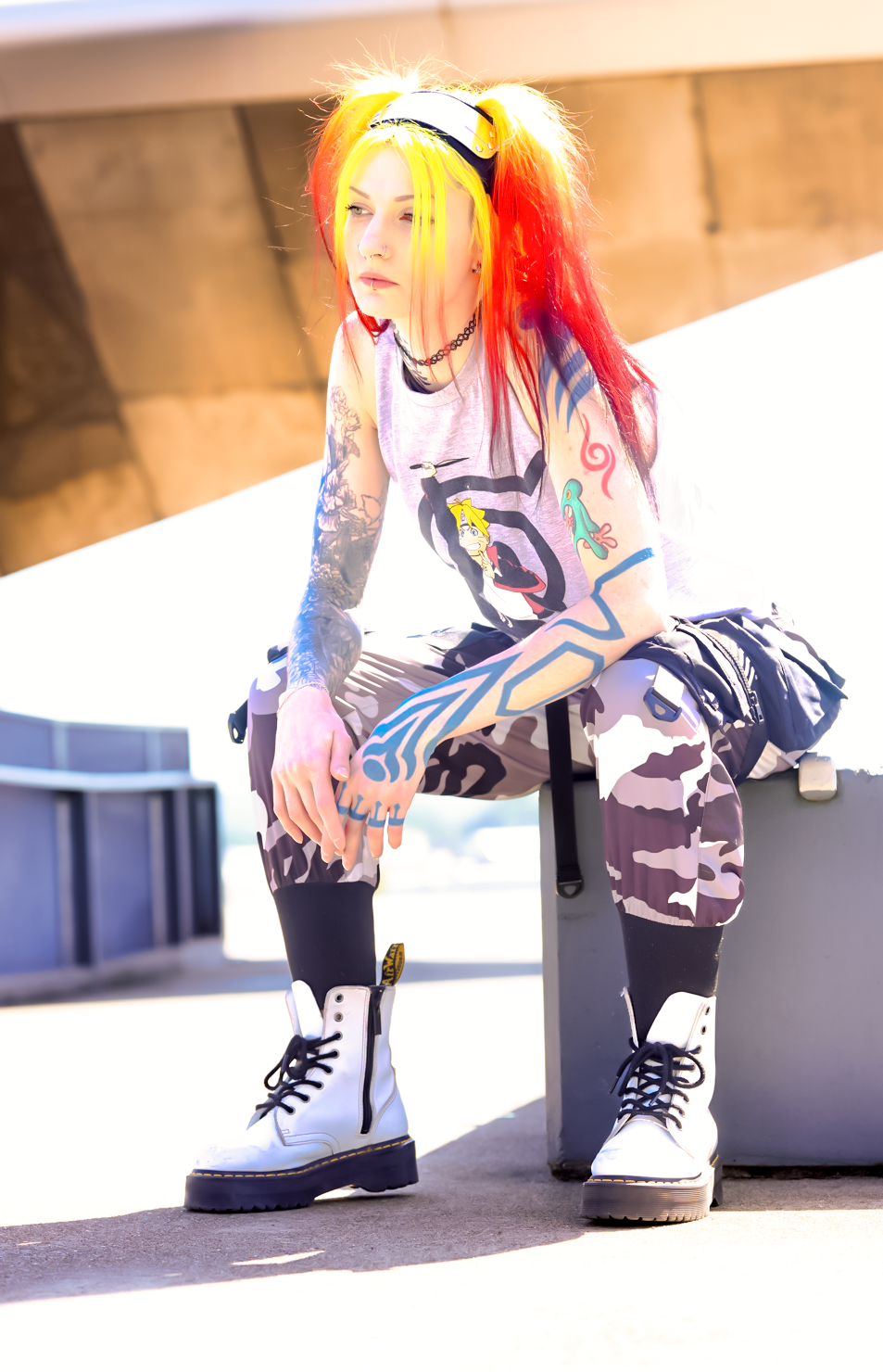 A person with vibrant red and yellow hair wearing a graphic t-shirt, camo pants and white combat boots poses in sunlight.