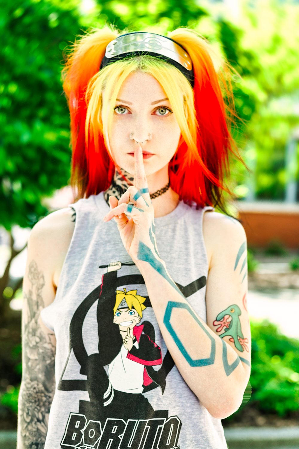 Anime cosplayer in Boruto shirt with red and yellow hair making shushing gesture against green outdoor background.