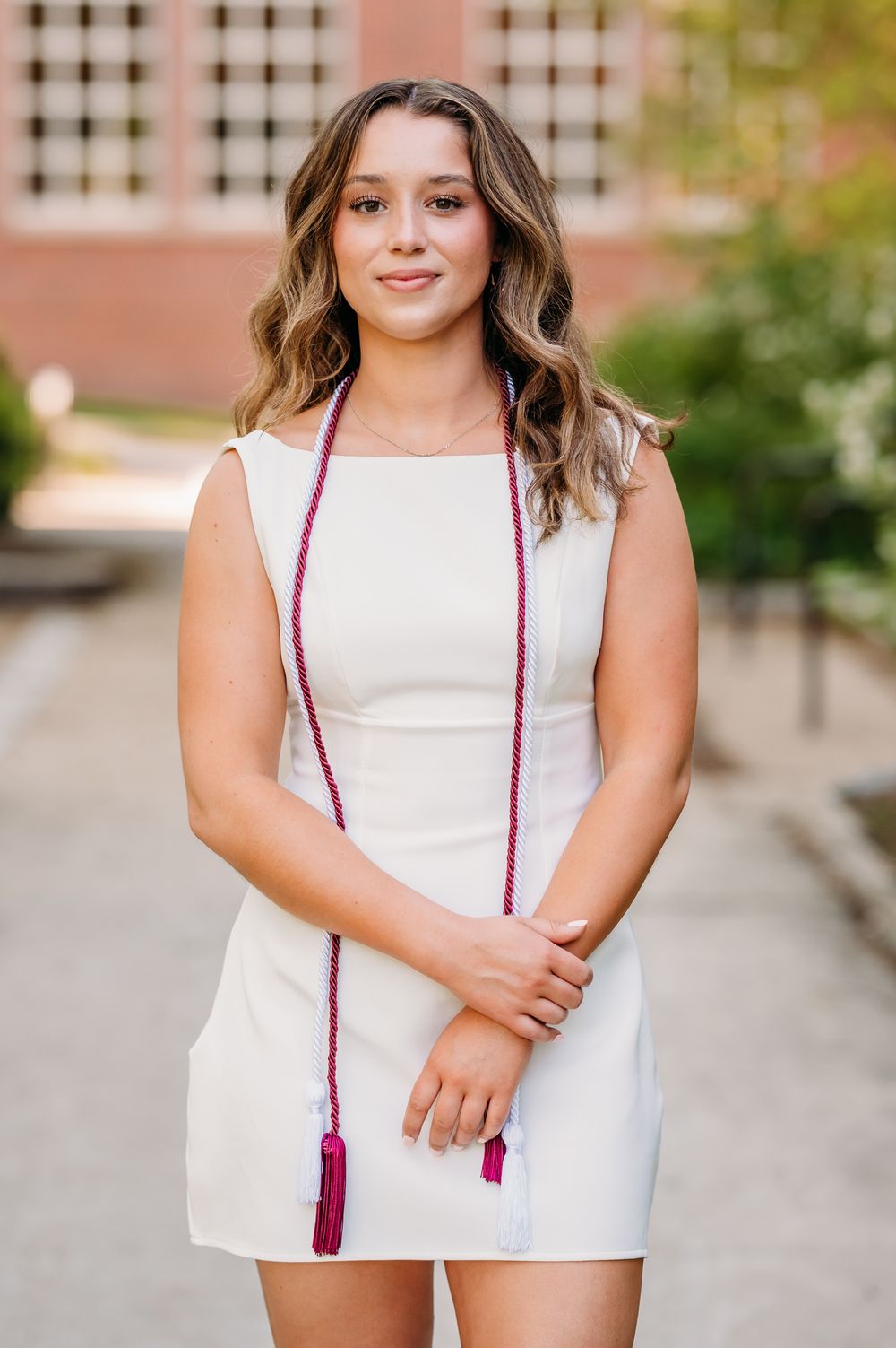 A person in a white sleeveless dress with wavy hair and red graduation cords stands outdoors on a sunny day.
