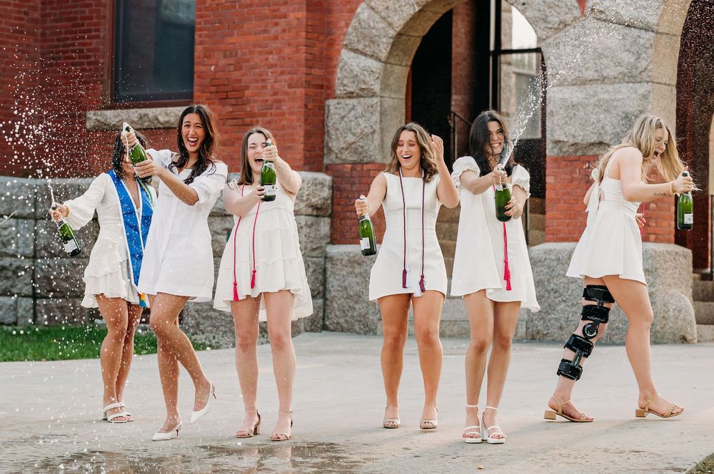 Group of friends in matching white dresses walk together laughing and smiling outside a brick building.