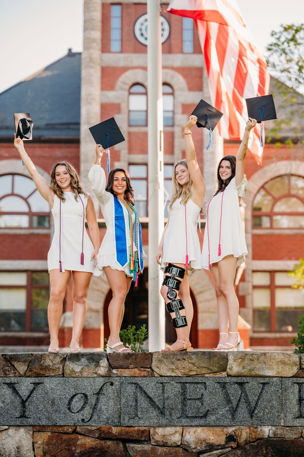 College graduates in white dresses celebrate graduation day by raising caps on historic university stone wall with American flag.