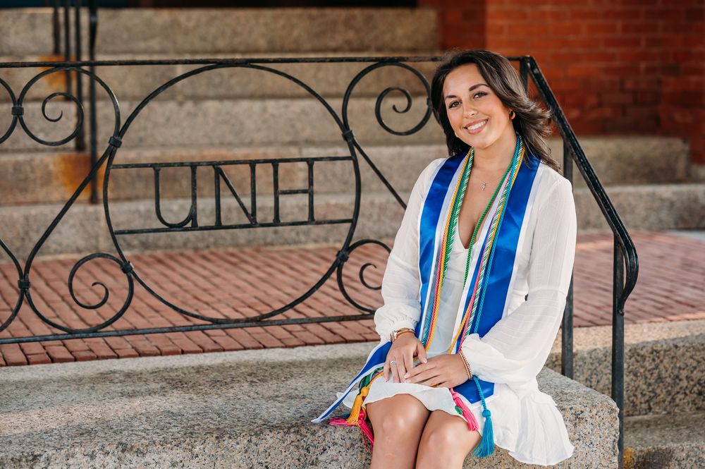 Graduate in white dress sitting on brick steps with ornate iron railing displaying university logo at commencement.