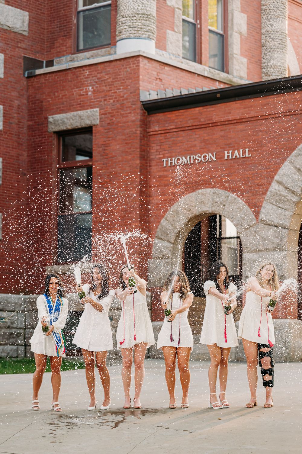 Group in white dresses celebrating outside Thompson Hall with water splashing in the air on a sunny day.