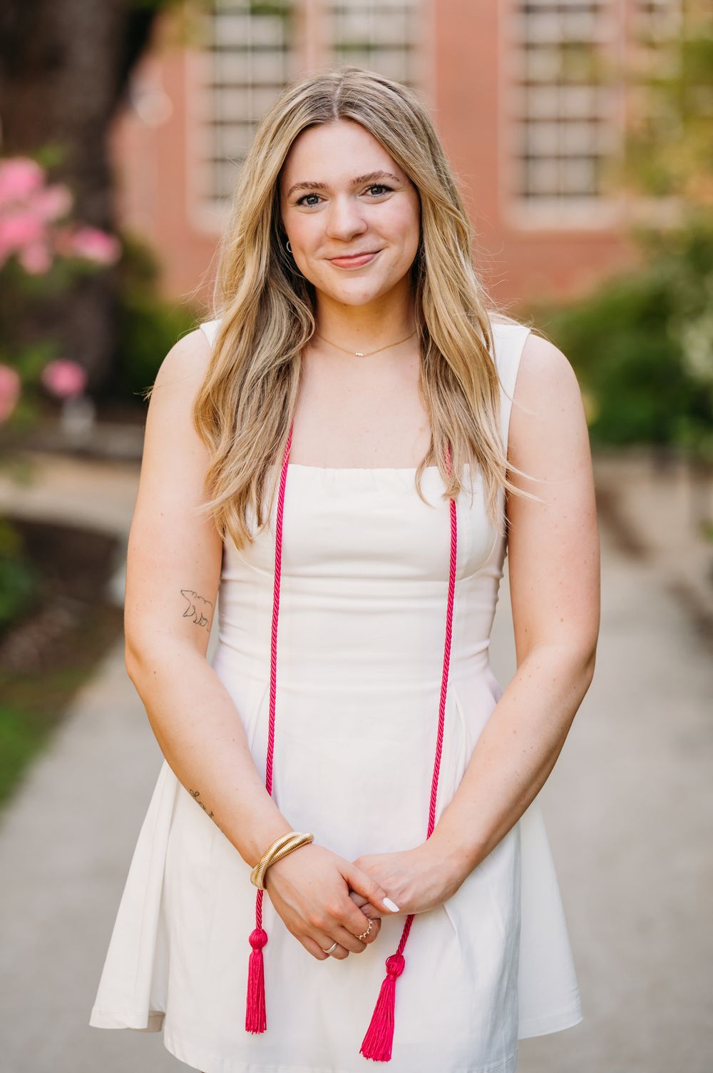 A person in a white sundress with red tassels stands in front of a brick building on a sunny day.