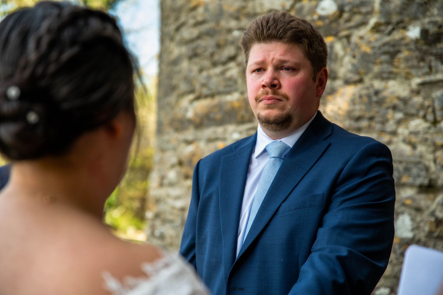 A groom in a navy blue suit listens intently during an outdoor wedding ceremony with a stone wall backdrop.