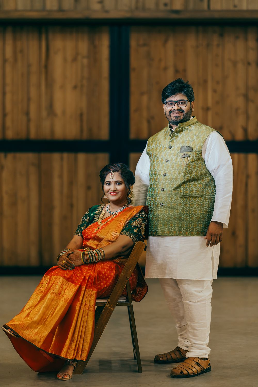 Telugu couple in traditional Indian attire poses together in front of a wooden wall, with the woman seated in a bright orange sari.