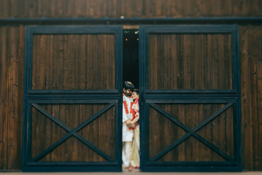 Telugu couple shoot through a gap between two large dark blue barn doors with X patterns against a wooden wall.