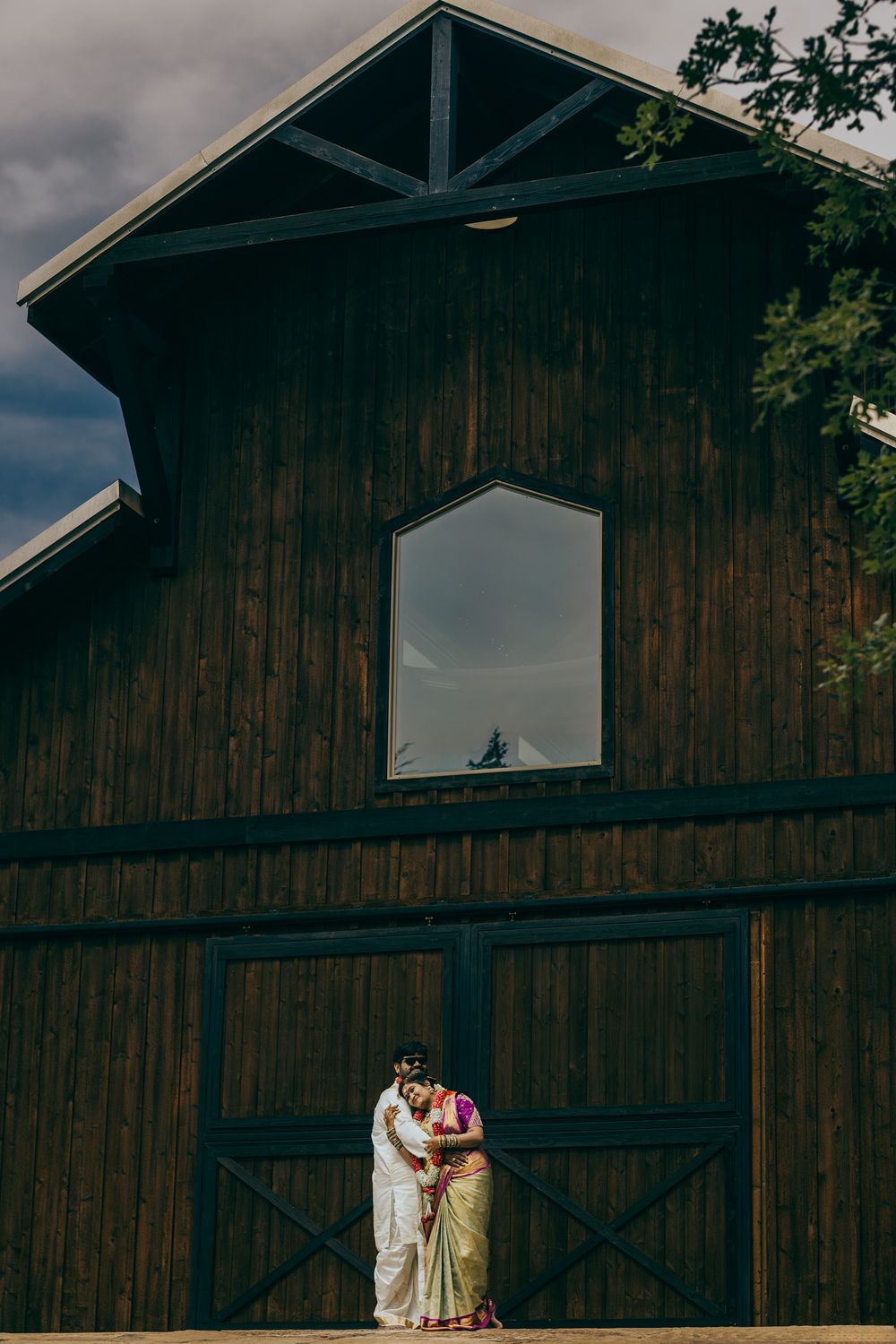 Telugu newly wedded couple pose in front of a rustic wooden barn with a white window under moody skies.