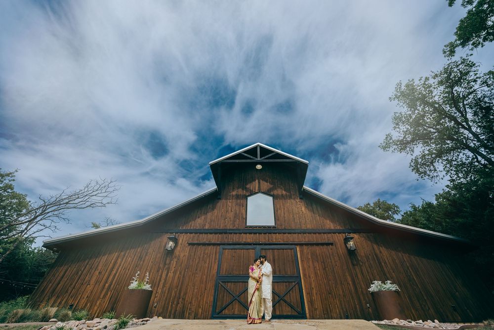 Telugu newly wedded couple pose in front of a rustic wooden barn with white accents under dramatic cloudy skies.