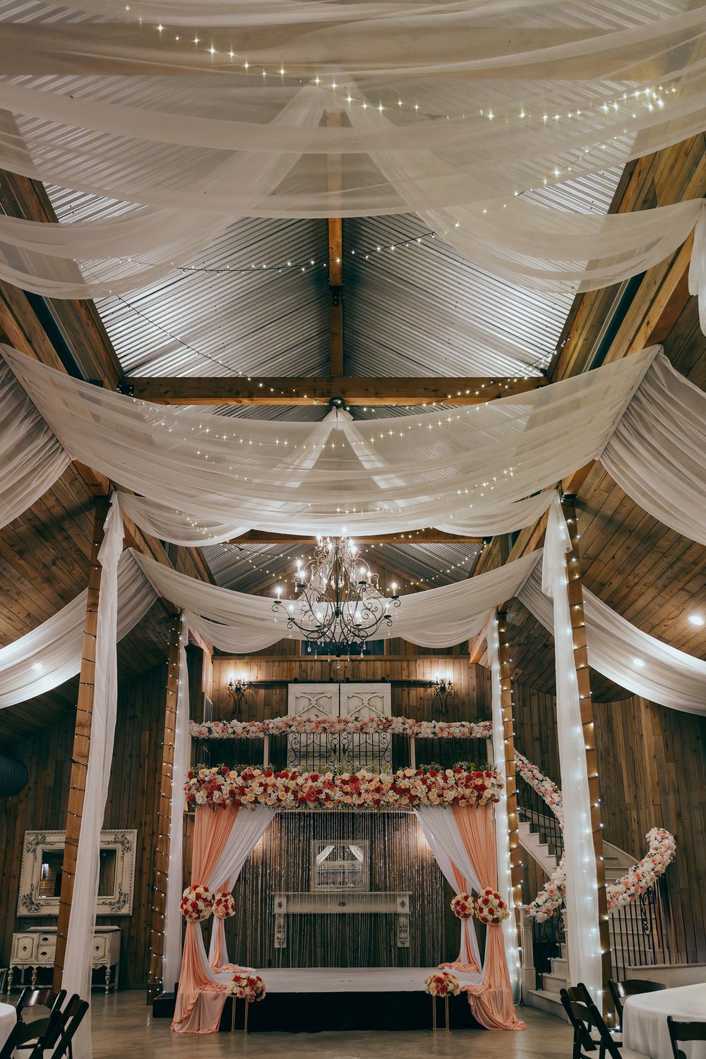Elegant wedding venue interior with white fabric draping and crystal chandeliers in a rustic barn setting.