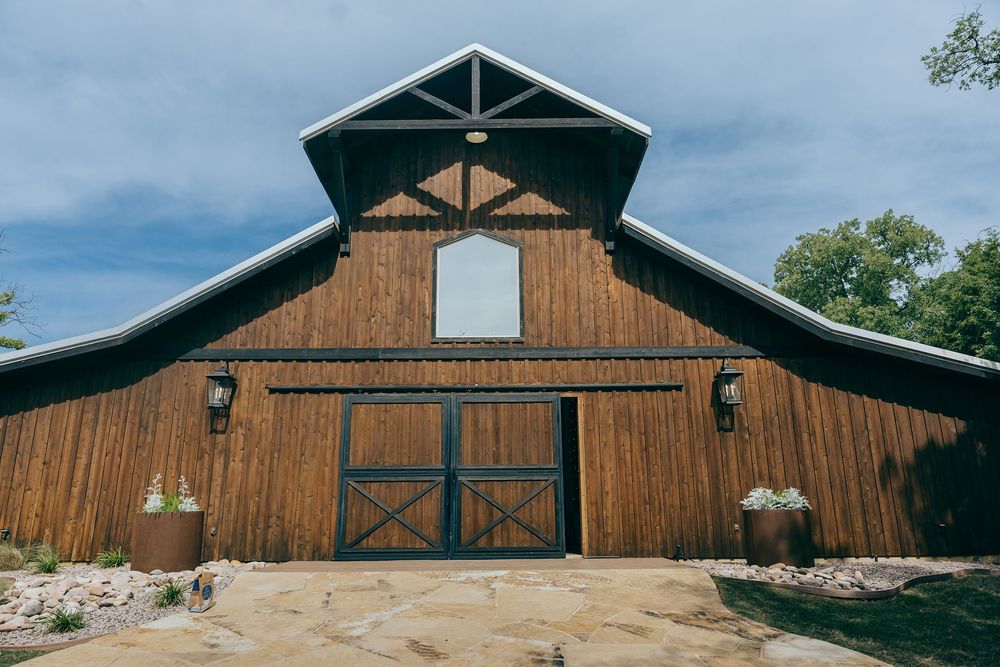A rustic wooden barn-style wedding venue with large doors and decorative gable features against a cloudy blue sky.