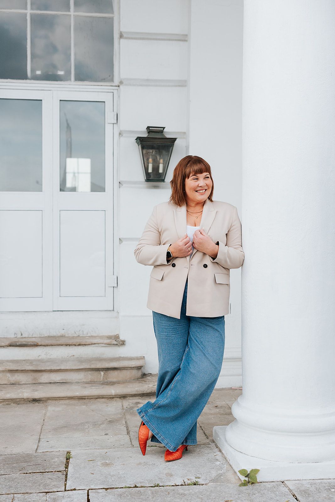 Person in beige blazer and blue jeans leans against white column while smiling outdoors.