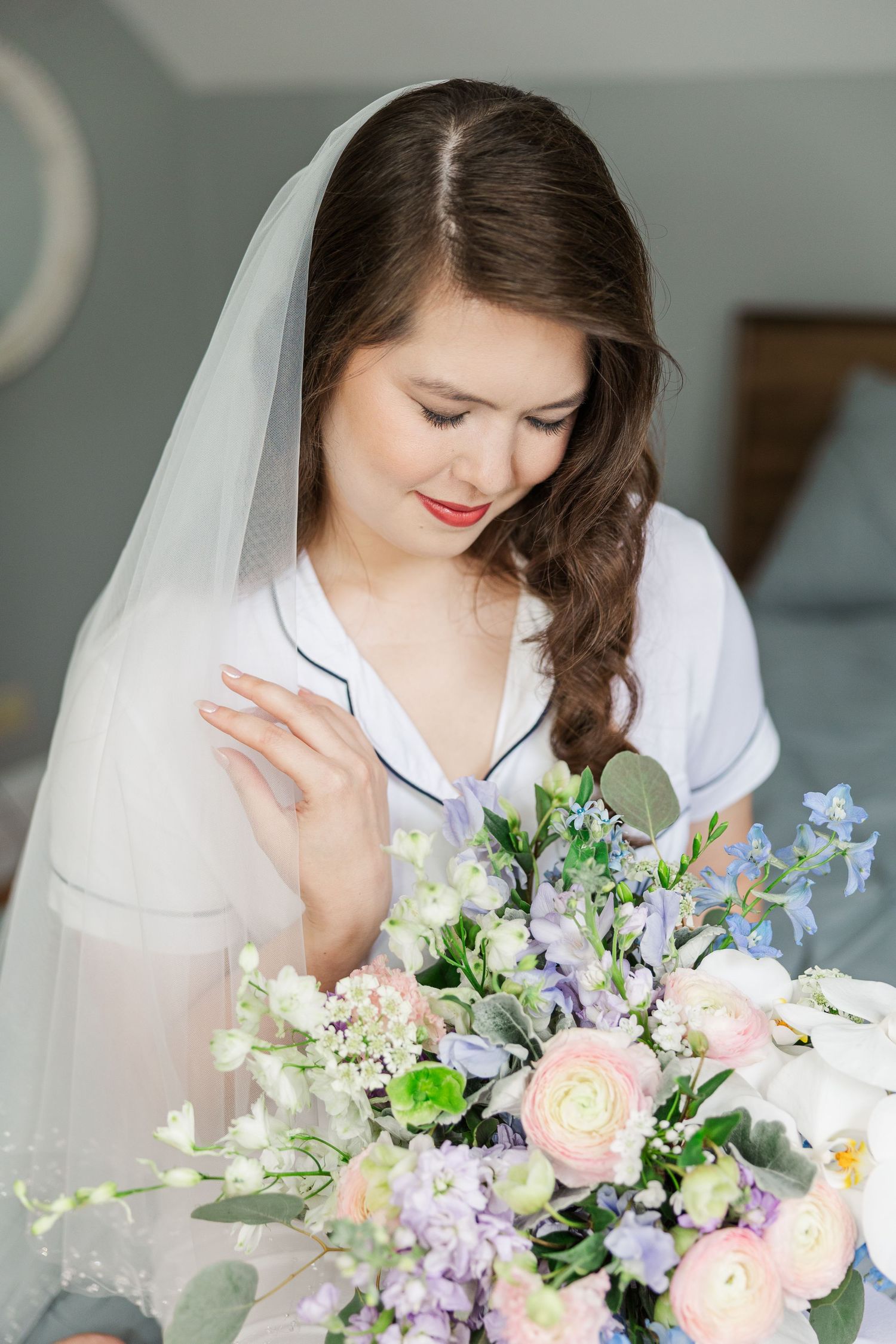 A bride in a white robe sits with her spring wedding bouquet featuring pink, purple and white flowers with eucalyptus.