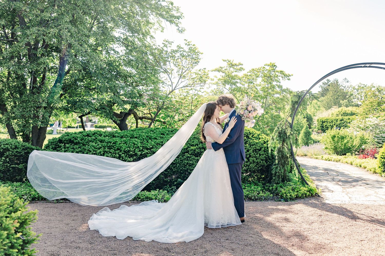 A couple in wedding attire embraces on a garden path while a long white veil flows dramatically in the breeze.