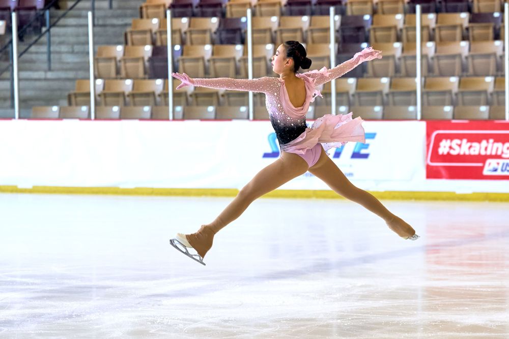 Figure skater performs a graceful split jump in a purple leotard on ice during competition performance.