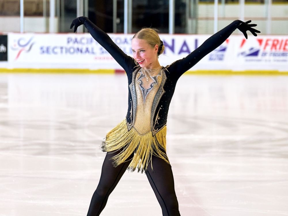 Figure skater performs on ice wearing black and gold costume with arms raised in graceful pose during competition.
