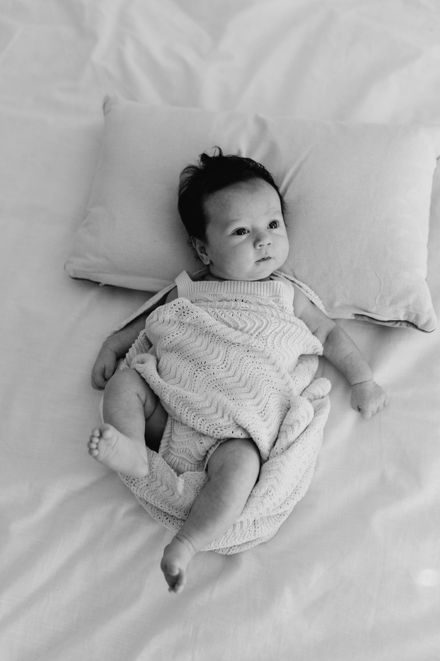 A baby wearing a white knitted outfit lies on crisp white bedding in a black and white photograph.