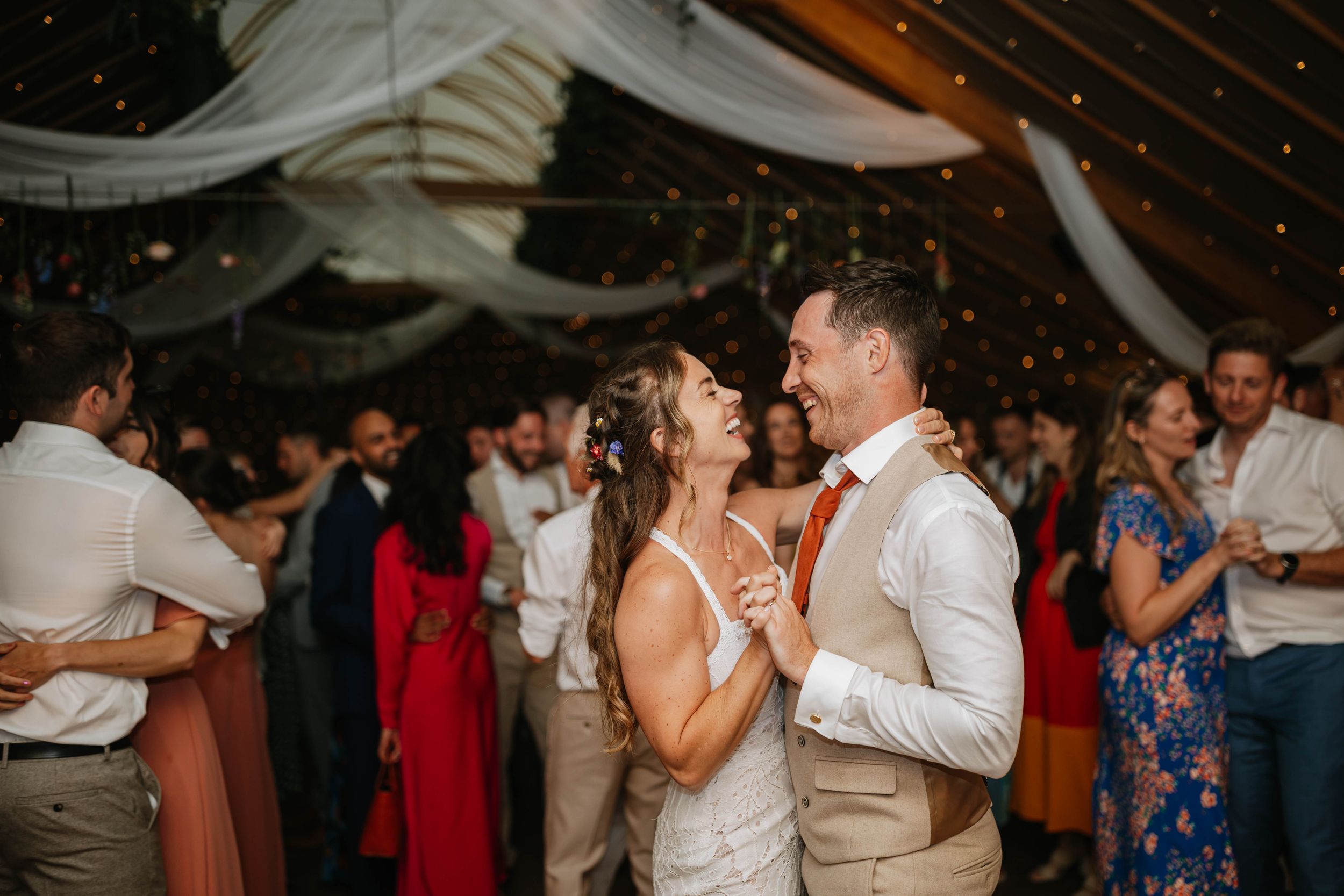 Couple shares a romantic slow dance at wedding reception under tent with soft lighting and guests in background.