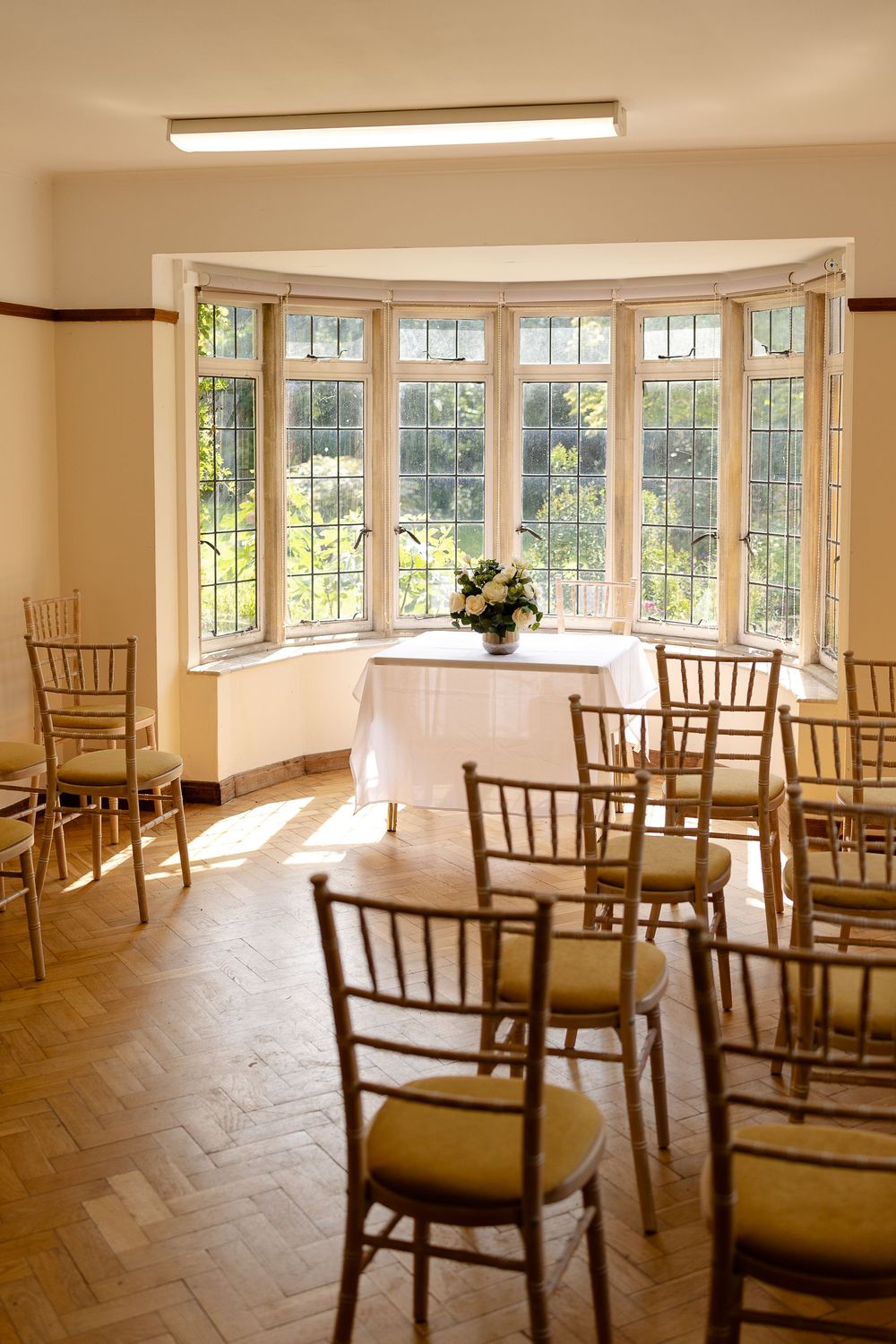 A bright ceremony room with rows of Chiavari chairs and a large bay window overlooking a garden.