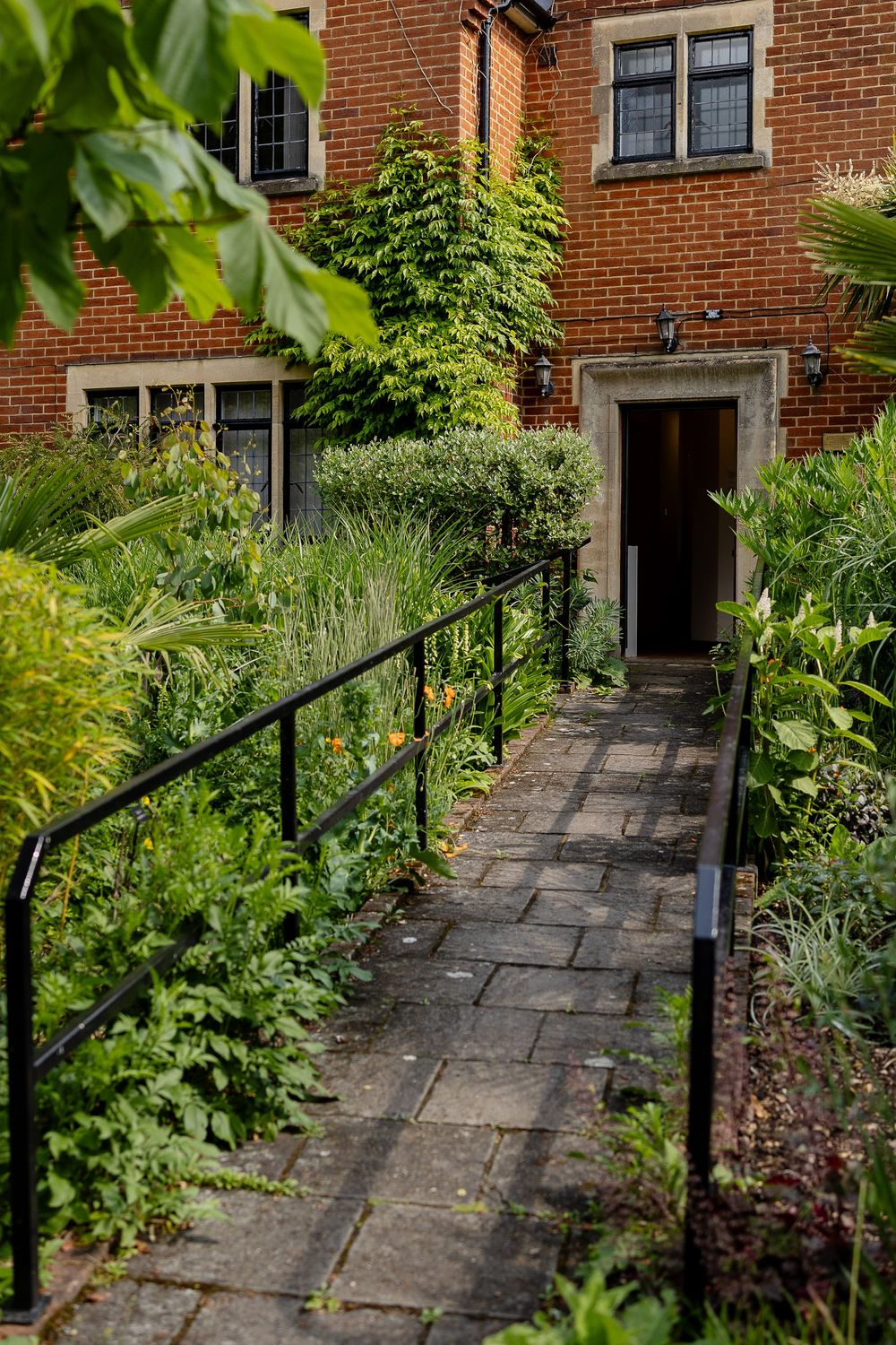 Stone pathway leading to a brick house entrance with lush garden borders and black metal handrails.