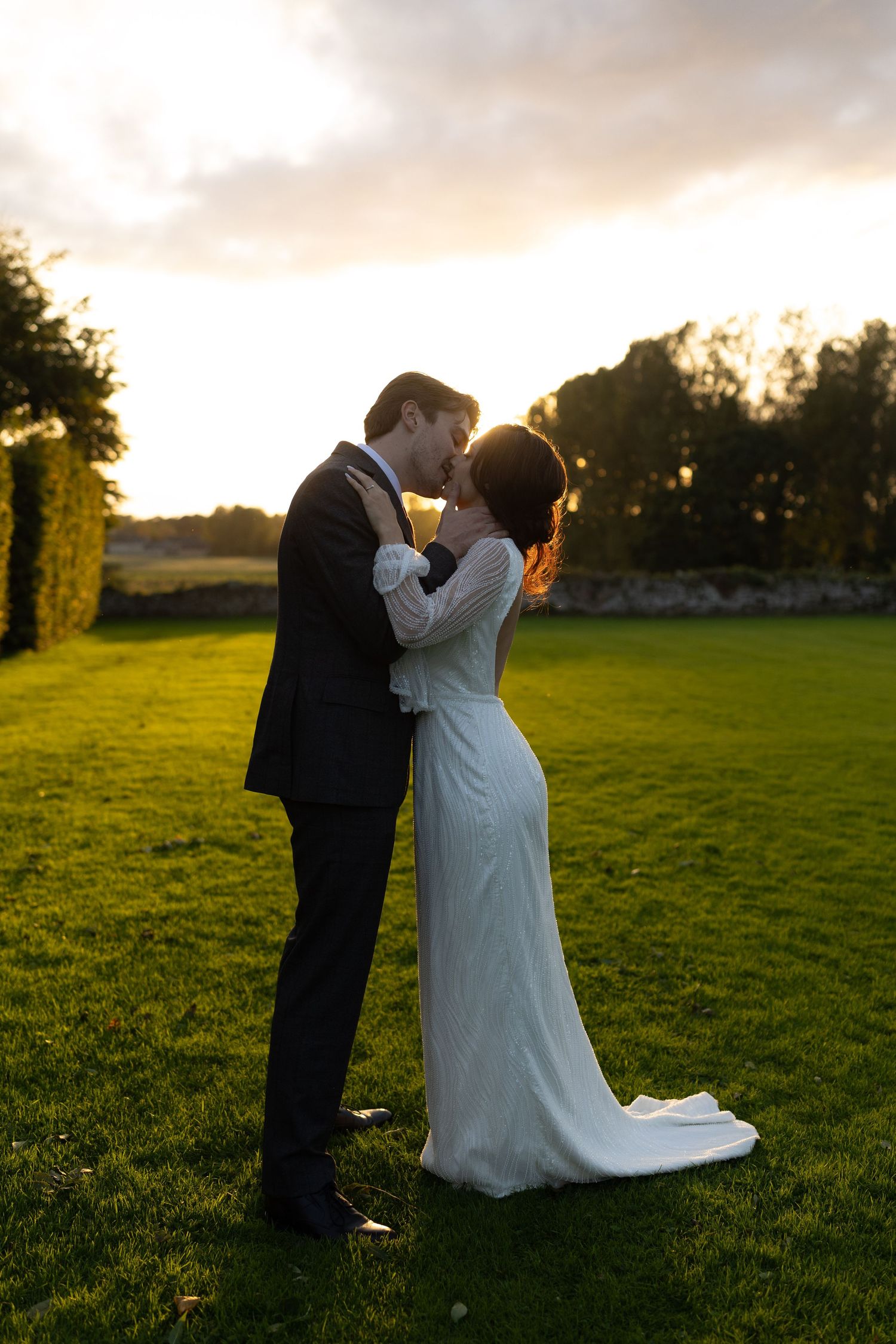 A couple in wedding attire embracing at sunset in a garden with golden light streaming through trees.