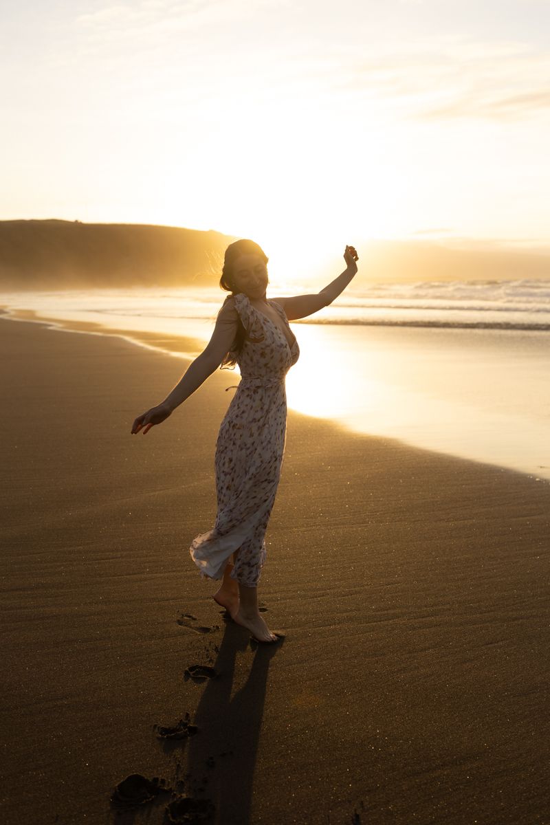 A person in a flowy dress twirls on a beach at sunset, casting a long shadow on the golden sand.