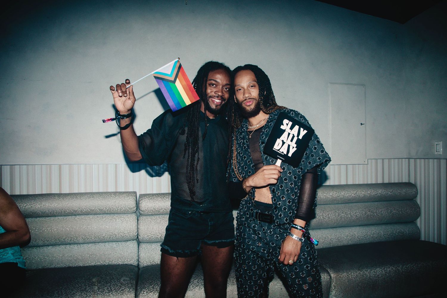 Two friends celebrate with pride flags at a nighttime event wearing black outfits and embracing while smiling.