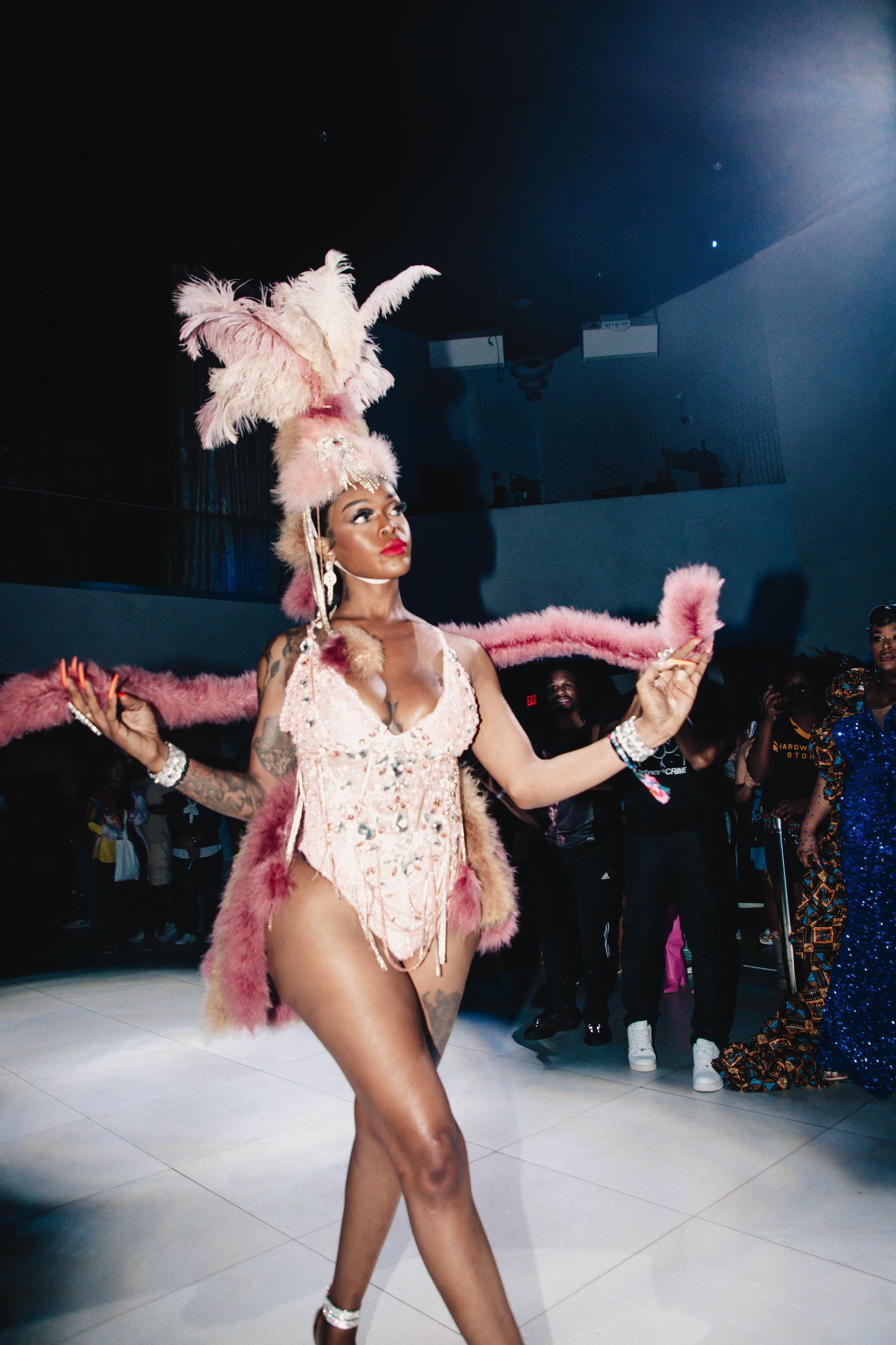 A performer in a white sequined leotard and pink feathers dances enthusiastically at a nightclub venue.