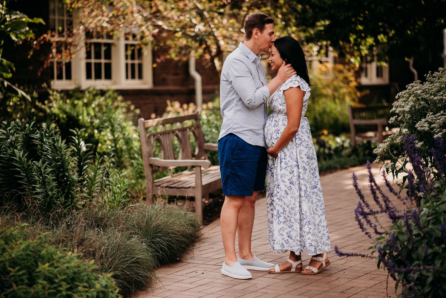 A couple shares a romantic moment together on a garden path surrounded by lush greenery and a brick building backdrop.